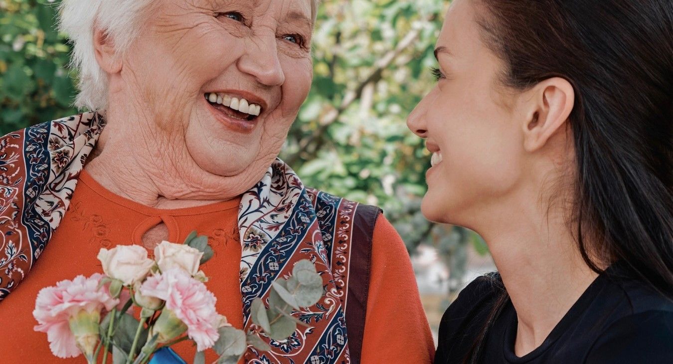 An elderly woman with a bouquet of carnations laughing together with a young volunteer